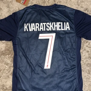PSG Kvaratskhelia #7 Nike matchtröja - Säljer en officiell Paris Saint-Germain fotbollströja från Nike med Kvaratskhelia och nummer 7 på ryggen. Tröjan är marinblå med röda och vita detaljer, PSG-emblem på bröstet och Qatar Airways som sponsor. Tillverkad i lätt och ventilerande material.