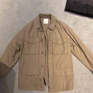 Beige overshirt från Dobber - Stilren beige overshirt från Dobber i bomull. Jackan har fyra stora fickor framtill med lock, klassisk krage och knäppning med knappar. Perfekt lager-på-lager-plagg med avslappnad passform och lång ärm.