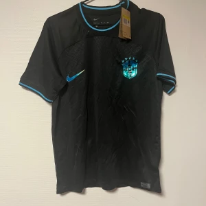 Svart Brasilien fotbollströja Nike S - Snygg svart Brasilien fotbollströja från Nike med blå detaljer och klubbmärke på bröstet. Tröjan är tillverkad i Dri-FIT-material som andas och har korta ärmar samt rund hals med blå rand. Perfekt för dig som vill sticka ut på planen eller på läktaren.