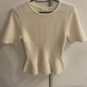 Vit stickad topp från H&M, S - Superfin vit stickad topp från H&M i storlek S. Modellen har korta ärmar, rund hals och en lätt utsvängd peplum-nederkant. Ribbad struktur ger en snygg och modern look. Perfekt att styla med jeans eller kjol för en fräsch vibe. HELT NY