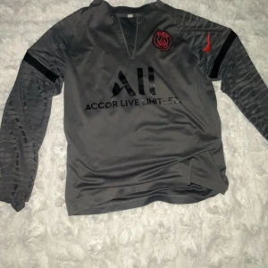 Grå långärmad PSG fotbollströja Nike - Snygg grå långärmad fotbollströja från PSG med svarta detaljer och röd Nike-logga. Tröjan har klubbmärke på bröstet, tryck med 'Accor Live Limitless' och 'Visit Rwanda' på ryggen. Tillverkad i ett glansigt, kedjan har gått av från tröjan. Strl S men passar Xs