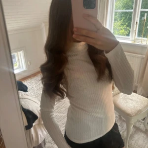 Vit ribbad polotröja - Säljer en vit ribbad polotröja med lång ärm och tight passform. Tröjan har en hög hals och är tillverkad i ett mjukt, stretchigt material som sitter snyggt på kroppen. Perfekt att styla med jeans eller kjol för en clean look.
