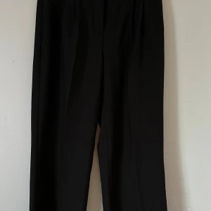 Svarta kostymbyxor från Bik Bok - Svarta kostymbyxor från Bik Bok i modellen PW Drew Tall. Byxorna har hög midja, raka ben och klassiska pressveck. Snygga bakfickor med knappar och bälteshällor upptill. Perfekta för en clean och stilren look.