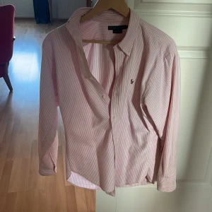 Rosa randig skjorta från Ralph Lauren - Säljer en klassisk rosa och vit randig skjorta från Ralph Lauren med broderad logga på bröstet. Skjortan har lång ärm, knappar framtill och en snygg krage. Perfekt för dig som gillar stilrena och tidlösa plagg.