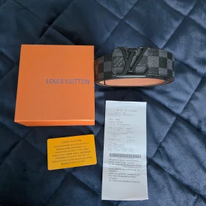 Svart Louis Vuitton Damier Graphite bälte - Snyggt Louis Vuitton bälte i klassiskt Damier Graphite-mönster med svart LV-spänne. Bältet är svart och grått i rutigt mönster och tillverkat i exklusivt skinn. Perfekt accessoar för att lyfta din outfit med lyxig känsla och ikonisk design.