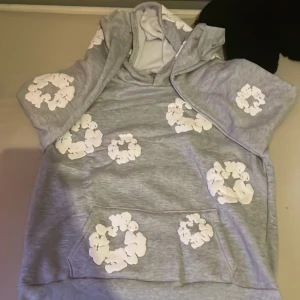 Grå hoodie med vita blommor från denim tears  - Grå hoodie med vita blommiga tryck över hela tröjan. Tröjan har huva med snörning och en stor magficka framtill. Långärmad och i mjukt material som känns skönt mot huden. Perfekt för chill dagar och när du vill ha en cool, avslappnad stil. Aldrig använd 