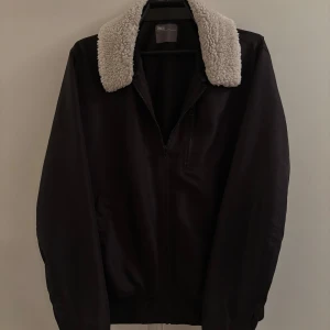 Svart teddyjacka från ASOS med krage - Passar strl M! Svart teddyjacka från ASOS i storlek L med fet, fluffig vit teddykrage. Jackan har dragkedja framtill och två sidofickor. Perfekt för dig som vill ha en stilren jacka med lite extra edge och mysig känsla.