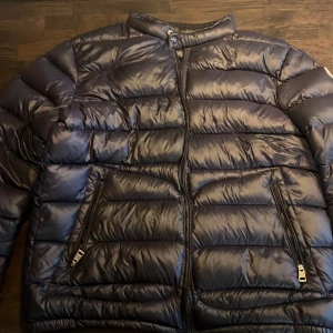 Mörkblå pufferjacka från Moncler - Säljer en mörkblåpufferjacka från Moncler Longue Saison med hög krage och två sidofickor med dragkedja. Jackan har ett stilrent, quiltat mönster och är fylld med dun för extra värme. Perfekt för kalla dagar och har en snygg, modern look den har ett litet hål vid armen och dragkedjan är lite trög därav de låga priset kom pm för mer bilder.