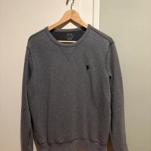 Grå sweatshirt från Polo Ralph Lauren - Snygg grå sweatshirt från Polo Ralph Lauren med klassisk rund hals och broderad svart logga på bröstet. Tröjan har långa ärmar och är tillverkad i mjuk bomull, perfekt för chill dagar. Enkel och stilren design som funkar till allt.