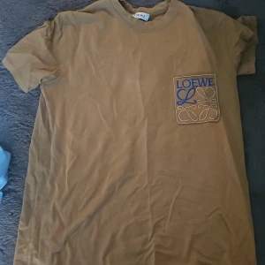 Beige Loewe t-shirt med broderad logga - Snygg beige t-shirt från Loewe med korta ärmar och rund hals. På bröstet finns en broderad patch med Loewe-loggan i blått och beige. Tillverkad i mjuk bomull för en skön känsla. Perfekt för dig som gillar stilrena och exklusiva plagg.