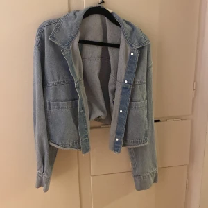 Ljusblå croppad jeansjacka M - Säljer en ljusblå croppad jeansjacka i klassisk denim med stora fickor framtill och knappar längs öppningen. Jackan har en avslappnad passform, bred krage och långa ärmar med knapp vid ärmslut. Perfekt för dig som gillar en chill och trendig stil.