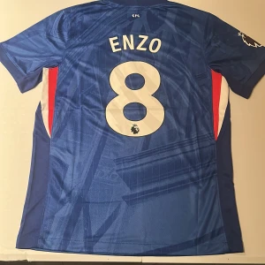 Chelsea Enzo #8 Nike matchtröja - Chelsea FC hemmatröja med Enzo och nummer 8 på ryggen. Tröjan är blå med vita och röda detaljer, har korta ärmar och är tillverkad i lätt, ventilerande polyester. Nike-logga och Chelsea-märke framtill, Premier League-märke på ärmen. Replika, endast handtvätt gäller!