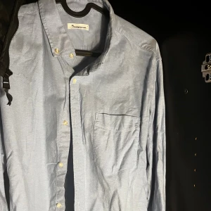 Ljusblå skjorta från Knowledge Cotton - Säljer en stilren ljusblå skjorta från Knowledge Cotton Apparel i storlek L. Skjortan har klassisk krage med knappar, bröstficka och långa ärmar. Tillverkad i mjuk bomull