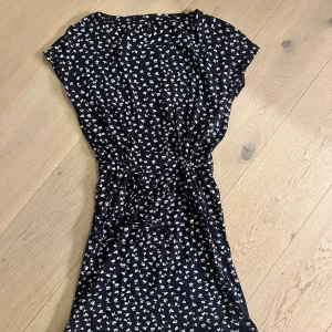 Svart blommig kortklänning med knyt - Supersöt svart kortklänning med små vita blommor och knyt i midjan. Klänningen har korta ärmar och en avslappnad passform som ger en skön och trendig look. Perfekt för varma dagar när du vill känna dig både bekväm och snygg.