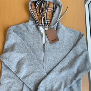 Grå hoodie från Burberry med dragkedja - Snygg grå hoodie från Burberry med klassiskt rutigt foder i huvan och längs dragkedjan. Tröjan har hel dragkedja, snörning i huvan och broderad logga på bröstet. Perfekt för dig som gillar stilrena och exklusiva plagg.