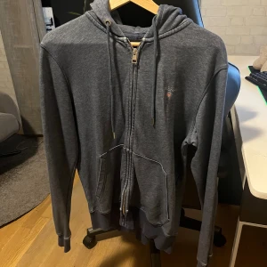 Mörkgrå hoodie från Gant med dragkedja - Mörkgrå hoodie från Gant med dragkedja framtill, snörning i huvan och två stora fickor. Klassisk broderad Gant-logga på bröstet. Perfekt för en avslappnad stil och enkel att matcha med jeans eller joggers.