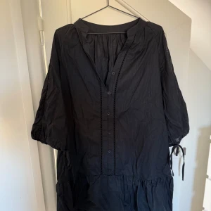 Svart kort klänning med volang - Svart kort klänning i bomull med knappar framtill, trekvartsärmar med knyt och volang nertill. Lätt och luftig modell med fina spetsdetaljer längs knappraden och ärmarna. Perfekt för dig som gillar en avslappnad men ändå stilren look samt att den aldrig är använd!🥰