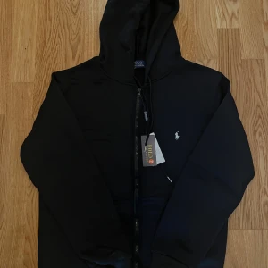 Svart zip-hoodie från Polo Ralph Lauren - Svart hoodie från Polo Ralph Lauren med dragkedja framtill och klassisk huva. Tröjan har det ikoniska broderade logotypen på bröstet och snörning vid huvan. 