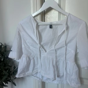 Vit blus med knyt från H&M Divided XS - Supersöt vit blus från H&M Divided i storlek XS. Blusen har v-ringning med knytband framtill, volang nedtill och halvlånga ärmar. Tillverkad i bomull för en luftig känsla. Perfekt till jeans eller kjol för en fräsch look.