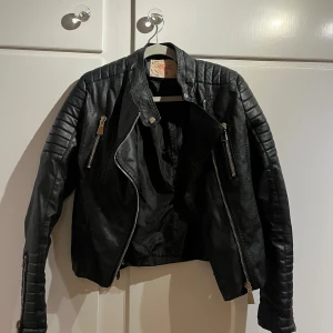 Svart skinnjacka från Voyelles, stl 38 - Snygg svart skinnjacka från Voyelles i bikerstil med quiltade detaljer på axlar och ärmar. Jackan har dragkedjor framtill och vid ärmsluten samt spännen i sidorna. Materialet är polyuretan med foder i polyester. Perfekt för en edgy look.