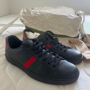 Gucci ace black leather  - Gucci Ace sneakers i svart skinn med klassiska gröna och röda ränder på sidorna. Baksidan har en röd, ormskinnsmönstrad detalj med Gucci-logga. Platt sula, rund tå och svarta skosnören. Snygga och ikoniska sneakers som lyfter varje outfit. Storlek:46 , Skick: Helt nya alldrig använda(bara provade), Ingår: orginalkartong, dust bags samt papper 