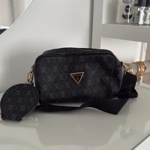 Svart axelväska från Guess med mönster - Snygg svart axelväska från Guess med justerbar rem och guldfärgade detaljer. Väskan har ett diskret mönster med loggor över hela ytan och en praktisk liten rund ficka på sidan. Tillverkad i syntetmaterial och stängs med dragkedja.