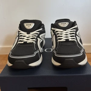Dior B30 svarta och vita sneakers - Säljer ett par Dior B30 sneakers i svart och vitt med stora CD-detaljer på sidorna. Skorna har en sportig siluett, mesh och syntetpaneler, samt vita snören och en vit sula. 