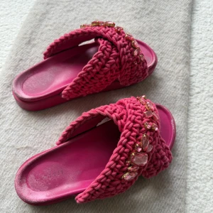 Inuikki Rosa sandaler med stickade band och stenar - Unika rosa sandaler med breda, stickade band i kors och stora rosa stenar som dekoration. Sulan är platt och matchar färgen på banden. Perfekta för dig som vill sticka ut med färg och detaljer. Superfina till sommarens outfits! Storlek 40 och nypris 2700kr
