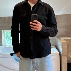 Svart overshirt i ull med fickor - Säljer en svart overshirt i ull med klassisk krage och två bröstfickor med lock och knappar. Skjortan har lång ärm, knäppning framtill och en clean, stilren look. Perfekt att slänga över en t-shirt för en chill vibe. Nypris 2100kr 