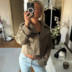 Grön trenchcoat  - Säljer en grön trenchcoat med bred krage, dubbelknäppning och bälte vid ärmsluten. Jackan har en boxig passform och stora knappar framtill. Perfekt för dig som vill ha en trendig och stilren look till våren eller hösten.
