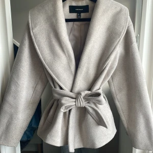 Beige omlottjacka från Vero Moda - Snygg beige jacka från Vero Moda med bred krage och knytskärp i midjan. Jackan har en omlottmodell, långa ärmar och praktiska fickor på sidorna. Perfekt för dig som gillar stilrena och trendiga plagg med en chill vibe.