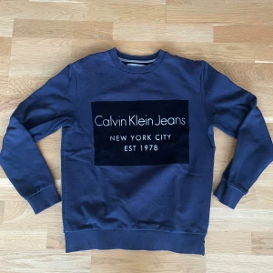 Blå sweatshirt från Calvin Klein Jeans - Mörkblå sweatshirt från Calvin Klein Jeans med stor logga framtill. Tröjan har rund hals, långa ärmar och ribbade muddar vid ärmslut och nederkant. Tillverkad i mjuk bomullsmix som känns skön mot huden.