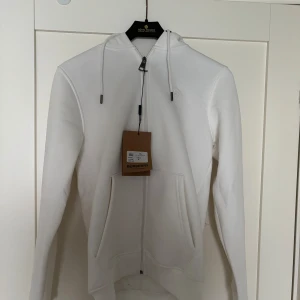 Vit hoodie från Burberry med dragkedja - Säljer en stilren vit hoodie från Burberry i bomull med klassiskt rutigt foder i huvan. Tröjan har hel dragkedja, snörning i huvan och två stora fickor framtill. Perfekt för dig som vill ha en clean look med en touch av lyx.