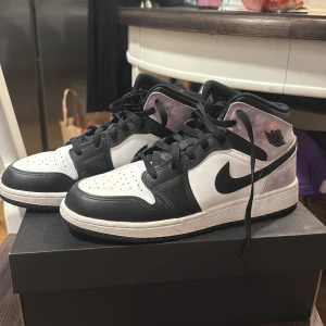 AIR JORDAN 1 MID SE. BLACK/BLEAChED CORAL-WHITE - Nike Air Jordan 1 Mid sneakers med svart och vit läderbas, lila och rosa toner på hälen och kragen. Klassisk svart swoosh, snörning och perforerad tå. Mjukt textilmaterial på bakre panelerna och ikonisk Jordan-logga på sidan. Har använt dem 2 gånger. 