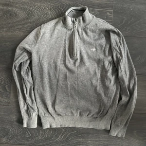 Grå half zip tröja från GANT - Grå långärmad tröja från GANT med halv dragkedja och broderad logga på bröstet. Tröjan har ribbade muddar och krage, perfekt för en clean och stilren look. Materialet är mjukt och bekvämt, passar till många outfits.ord pris 1099 knappt använt den 