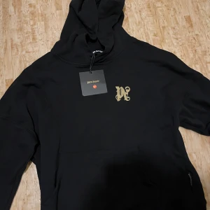 Svart hoodie från Palm Angels S - Svart hoodie från Palm Angels med stor broderad logga och drakmotiv på ryggen samt mindre logga framtill. Klassisk huva och känguruficka. Tillverkad i mjuk bomullsblandning med polyester. Grym streetwear-vibe och snygga detaljer.