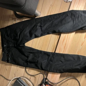 Svarta Levis 501 jeans med slitningar - Svarta Levis 501 jeans med snygga slitningar på benen. Klassisk rak passform och femficksmodell. Jeansen har knappar i gylfen och är tillverkade i robust denim. Perfekta för dig som gillar en lite mer edgy stil. 