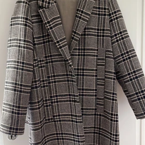 Rutig kappa från Zara TRF, storlek L - Säljer en snygg rutig kappa från Zara TRF Collection i svart och beige. Klassisk oversized modell med bred krage och knappar framtill. Perfekt för dig som gillar statement-plagg med coolt mönster och vill ha en jacka som sticker ut.