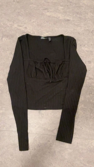 Svart ribbad långärmad topp med knyt - Svart croppad topp med långa ärmar och ribbad struktur. Toppen har en fyrkantig urringning och en söt knytning framtill under bysten. Perfekt för dig som gillar en stilren men ändå trendig look.