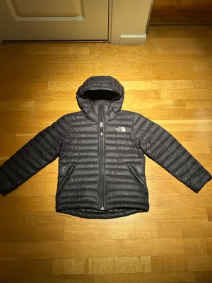 Svart dunjacka från The North Face - Säljer en svart dunjacka från The North Face med huva och dragkedja. Jackan är quiltad och har två fickor framtill. Perfekt för kalla dagar och har en clean, sportig look med logga på bröstet. Jackan passar 140-150 cm. Original pris 2500kr. Säljs för endast 399kr.