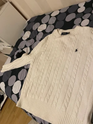 Vit kabelstickad tröja Polo Ralph Lauren - Vit kabelstickad tröja från Polo Ralph Lauren med klassisk rund hals och broderad marinblå logga på bröstet. Tröjan har ribbade muddar och är långärmad, perfekt för dig som gillar stilrena och tidlösa plagg.