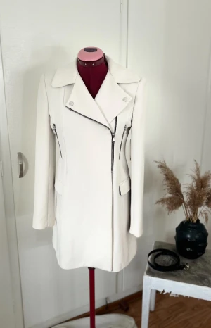 Vit rock med dragkedjor och krage - Snygg vit rock med klassisk krage och coola silverfärgade dragkedjor framtill. Jackan har lång ärm, två fickor med lock och en stilren, rak passform. Materialet känns mjukt och har en lätt struktur, perfekt för dig som gillar minimalistisk och clean stil. Visar även olika sätt man kan ha den på sig både stängd och öppen(lägger den billigt för att kunna få sålt snabbt)