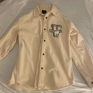 Beige overshirt i manchester - Snygg beige overshirt i manchester med coola broderade patches på bröstet och ärmen. Skjortan har långa ärmar, knappar framtill och en avslappnad passform. Perfekt för lager-på-lager och streetstyle.