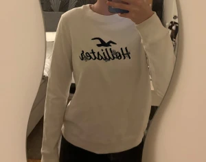 Vit Hollister sweatshirt med broderi - Vit sweatshirt från Hollister med mörkblå broderad logga och små blommor på bröstet. Tröjan har rund hals, ribbade muddar och är långärmad. Mjuk insida i fleece och klassisk passform 🩷