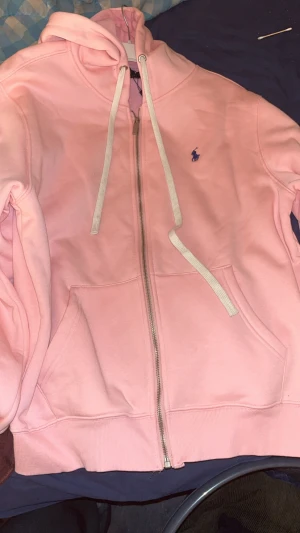 Rosa hoodie från Polo Ralph Lauren XL - Snygg rosa hoodie från Polo Ralph Lauren i storlek XL. Mjuk bomullsmix, dragkedja framtill, två fickor och vit dragsko i huvan. Klassisk blå Polo-logga broderad på bröstet och detaljerad zip med logotyp. Perfekt för lugnare  dagar. Kan tänka mig att sänka pris, köpt för runt 2000kr, inga defekter 