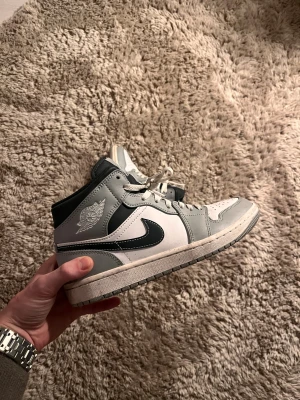 Nike Air Jordan 1 Mid grå/vit - Säljer ett par Nike Air Jordan 1 Mid i grått, vitt och svart. Klassisk siluett med svart swoosh, perforerad tåbox och snörning. Skorna har en mellanhög design och är tillverkade i skinn och syntet. Perfekta för dig som gillar streetwear och sneakers med ikonisk stil.