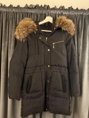 Svart dunjacka Hollies med fuskpäls - Snygg svart dunjacka från Hollies med stor fluffig fuskpäls på huvan, guldiga dragkedjor och knappar. Jackan har flera fickor, markerad midja och långa ärmar. Perfekt för kalla dagar när du vill vara både varm och stilren. Orginal pris 5000kr