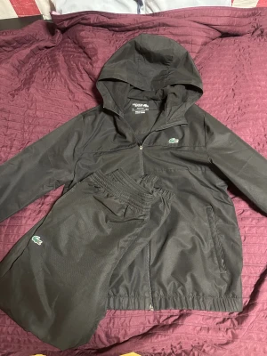 Svart tracksuit från Lacoste med huva - Svart tracksuit från Lacoste med klassisk krokodillogga på både jacka och byxa. Jackan har huva, dragkedja och två fickor. Byxorna har resår i midjan och vid benslut. Materialet är syntet och passformen är normal. Perfekt för chill eller sport. Jackan är M och byxorna är S men dem är relaxed fit så passar mer som M. Inga skador eller fläckar på dressen.