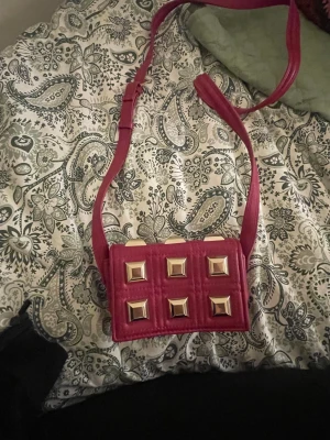 Rosa quiltad axelväska med nitar - Säljer en liten rosa väska från Zara med  guldiga fyrkantiga nitar på framsidan. Väskan har justerbart band. Zara märket i väskan har lossnat i en av sömmarna men inget som inte går att sy fast eller ta bort:) 