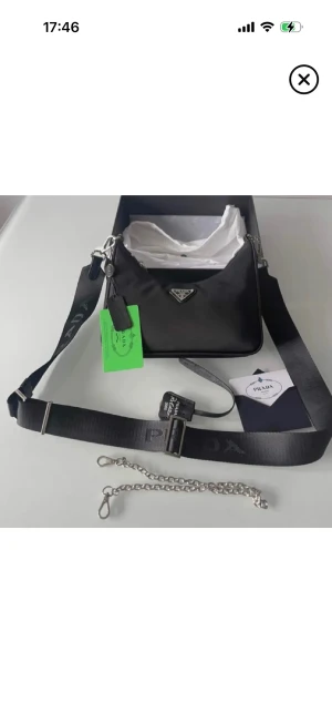 Svart axelväska från Prada  - Snygg svart axelväska från Prada i  med silverfärgade detaljer och justerbar axelrem. Väskan har klassisk trekantslogga framtill, dragkedja och medföljande liten pouch. Perfekt för dig som vill ha en stilren och trendig väska med lyxig känsla.